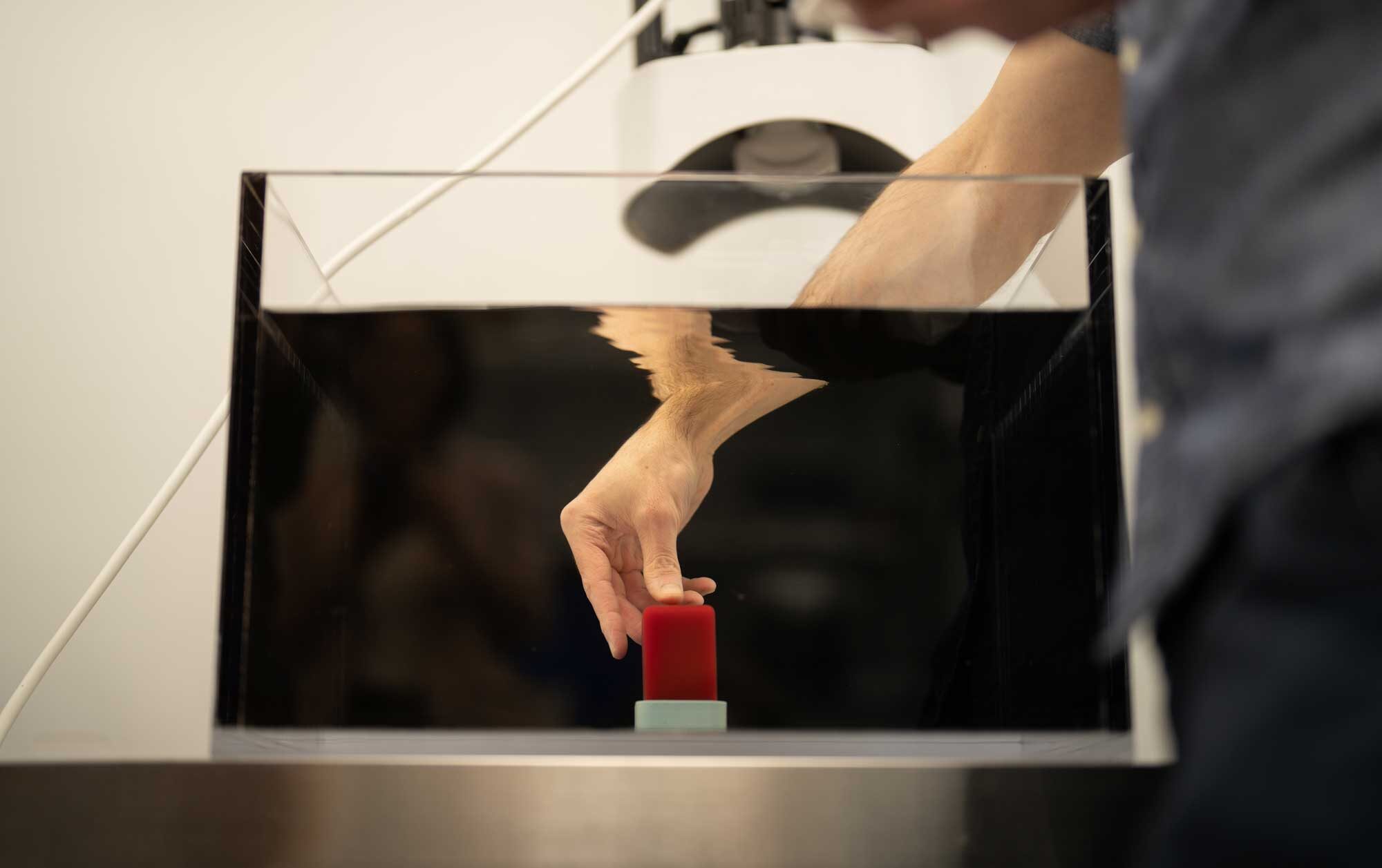 Duryea reaches his arm in a glass tank of water to adjust a red block at the bottom.