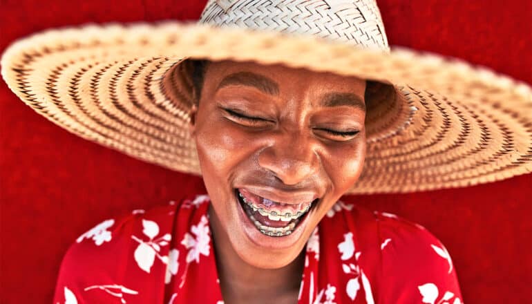 A woman wearing braces laughs and smiles widely while wearing a straw hat.